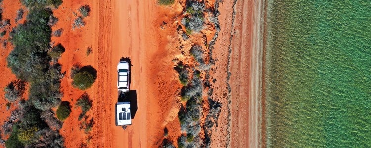 Aerial view of a camper on a red sandy road, bordered by vibrant greenery and a calm turquoise ocean on the right.