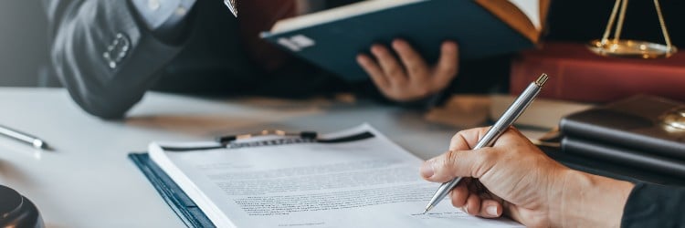 A close-up of a hand holding a silver pen, signing a document, while another person reads a book in a legal setting.
